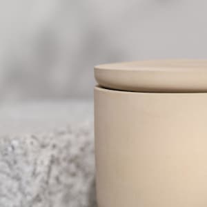 May include: A beige ceramic jar with a matching lid. The jar has a cylindrical shape with a slightly rounded base. The lid sits flush on top, creating a clean, minimalist aesthetic. The background is blurred, highlighting the jar's form.