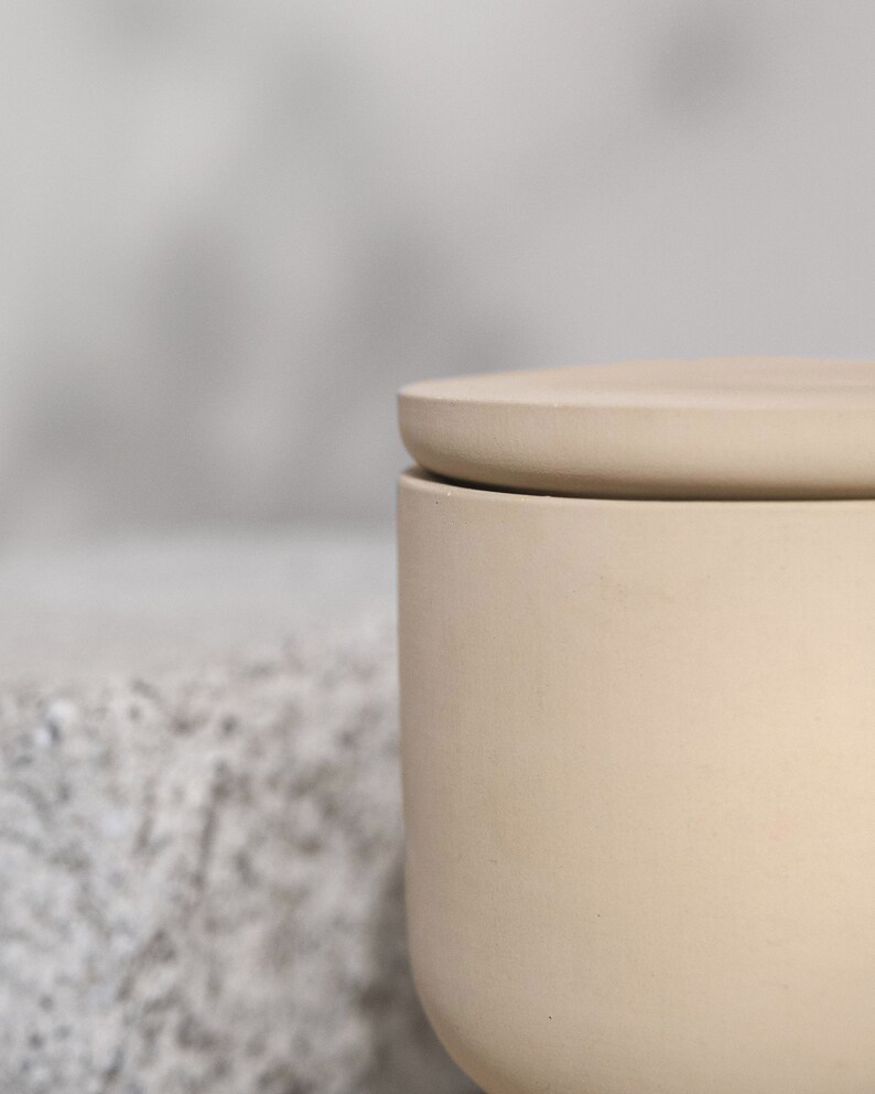 May include: A beige ceramic jar with a matching lid. The jar has a cylindrical shape with a slightly rounded base. The lid sits flush on top, creating a clean, minimalist aesthetic. The background is blurred, highlighting the jar's form.