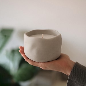 Pode incluir: Uma vela cinza artesanal em um recipiente de cerâmica com formato orgânico. A vela tem um interior de cera branca e um único pavio de madeira. O recipiente é segurado em uma mão, com um fundo neutro. É um item de decoração.