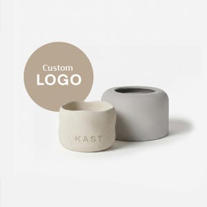 Può includere: Due fioriere minimaliste in cemento, una grigio chiaro e una bianco sporco, con la scritta "KAST" sulla fioriera bianco sporco. Un cerchio beige con la scritta "Custom LOGO" è sullo sfondo. Le fioriere hanno un design moderno e arrotondato.