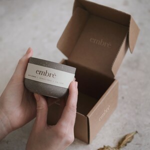 May include: A speckled gray candle in a cylindrical container with a white label that reads "embr&egrave;". The candle is held in front of an open cardboard box with the "embr&egrave;" logo. The box is also brown. The candle is 4 oz / 113 g.