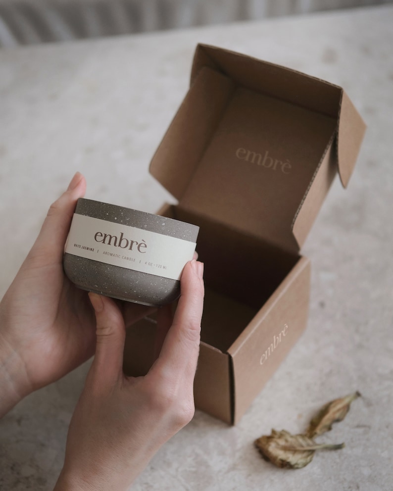 May include: A speckled gray candle in a cylindrical container with a white label that reads "embr&egrave;". The candle is held in front of an open cardboard box with the "embr&egrave;" logo. The box is also brown. The candle is 4 oz / 113 g.