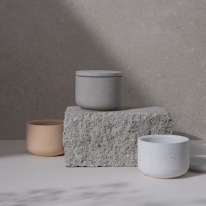 May include: Three minimalist ceramic containers in neutral tones. A light gray lidded jar sits atop a textured stone block, with a peach-colored pot and a speckled white pot nearby. The scene is lit with soft, natural light.