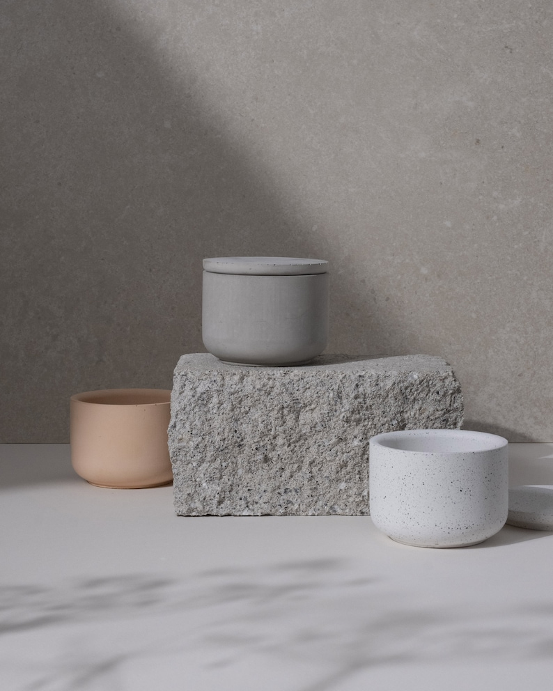 May include: Three minimalist ceramic containers in neutral tones. A light gray lidded jar sits atop a textured stone block, with a peach-colored pot and a speckled white pot nearby. The scene is lit with soft, natural light.