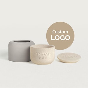 Può includere: Un set di tre contenitori in ceramica dai toni neutri. Un vaso cilindrico grigio, un vaso beige con il testo "ANOMI STUDIO" e un coperchio abbinato. Un cerchio marrone con "Custom LOGO" sullo sfondo.
