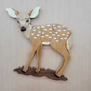 May include: Wooden wall art of a fawn with white spots on its light brown body. The fawn has large ears and dark eyes. It stands on a dark brown wooden base, creating a charming woodland scene. The artwork is made of various wood pieces.