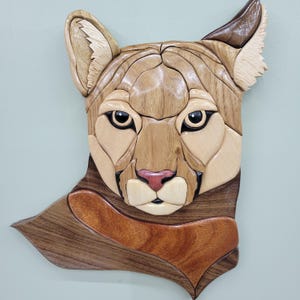 May include: A wall-mounted intarsia wood art piece depicting the head of a mountain lion. The sculpture is crafted from various wood tones, including light tan, medium brown, and dark walnut, with a small pink-toned wood accent on the nose.