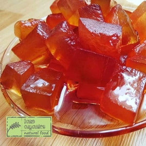 Op de afbeelding: Een glazen kom gevuld met doorschijnende rode gelei blokjes. De kom staat op een houten oppervlak. De tekst "take organics naturel food" is zichtbaar op een groene bladsticker.