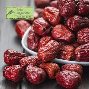 May include: A white bowl filled with dried red dates, with more dates scattered on a dark wooden surface. The bowl is on the right side of the image, and the dates are in focus. The text "take organics natural food" is on a green label in the upper left corner of the image.
