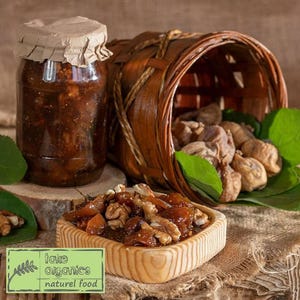 Peut inclure: Un pot en verre rempli de confiture de figues et de noix, avec un panier en osier brun rempli de figues séchées en arrière-plan. Le pot a un couvercle en papier brun et le texte "take organics naturel food" est sur une étiquette verte au premier plan.