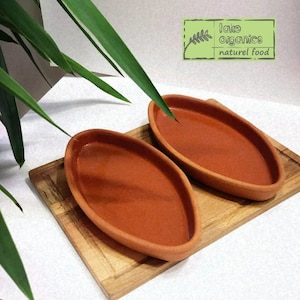 Könnte beinhalten: Zwei ovale Servierplatten aus Terrakotta auf einem Holzbrett. Die Platten sind rotbraun und haben eine leicht glasierte Oberfläche. Das Brett ist hellbraun und hat eine natürliche Holzmaserung.