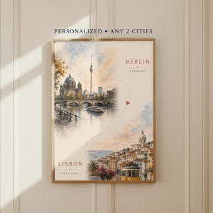 Op de afbeelding: Een ingelijste wandkunstprint met een aquarelillustratie van Berlijn, Duitsland, en Lissabon, Portugal. De tekst luidt PERSONALIZED ANY 2 CITIES, BERLIN GERMANY en LISBON PORTUGAL. Een dunne lijn met een klein hartje verbindt de twee stadsgezichten.