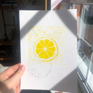 May include: A yellow lemon print on white paper. The lemon is in the center of the paper and is surrounded by a yellow frame.