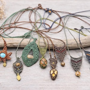 Könnte beinhalten: Eine Sammlung handgefertigter Makramee-Halsketten in verschiedenen Designs und Farben. Die Halsketten weisen aufwendige Knotenarbeiten, Edelsteine und Anhänger auf. Die Farbpalette umfasst Braun, Grün, Blau und Gold, wobei jede Halskette einen einzigartigen Stil hat.