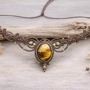Op de afbeelding: Een bruine macramé ketting met een ovale tijgeroogsteen in het midden. De ketting heeft ingewikkeld knoopwerk, kleine goudkleurige kralen en bloemvormige accenten. De ketting wordt gepresenteerd op een houten oppervlak.