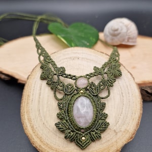 Puede incluir: Un collar de macramé verde con un gran colgante ovalado de cuarzo rosa y una pequeña cuenta redonda de cuarzo rosa. El collar se muestra sobre una superficie de madera.