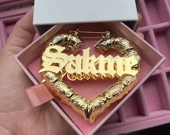 May include: Gold-tone heart-shaped hoop earrings with the name "Sakone" spelled out in bold, gothic-style lettering. The earrings are in a pink jewelry box.