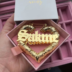 May include: Gold-tone heart-shaped hoop earrings with the name "Sakone" spelled out in bold, gothic-style lettering. The earrings are in a pink jewelry box.