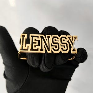 May include: A gold-toned metal belt buckle featuring the word LENSSY in a bold, block-style varsity font. The letters have a polished gold finish with black enamel detailing within the recessed areas of the lettering, creating a high-contrast look.