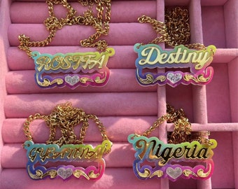 Könnte beinhalten: Vier goldene Halsketten mit bunten Namensschildern. Die Namensschilder haben einen Regenbogen-Verlauf und ein kleines Herz mit Glitzer. Die Namen auf den Namensschildern sind "Rosita", "Destiny", "Gianna" und "Nigeria".