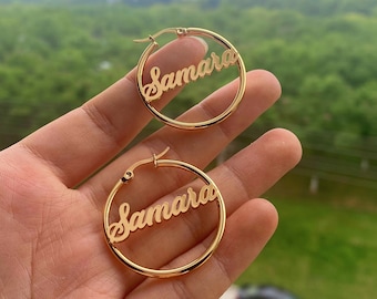 May include: Gold hoop earrings with the name "Samara" cut out in the metal. The earrings are shown on a hand against a blurry background.