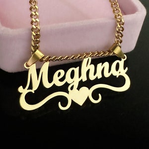May include: Gold necklace with a pendant that says "Meghna" in a cursive font with a heart at the bottom.