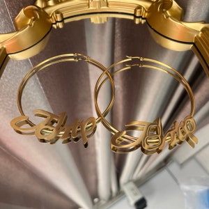 May include: A pair of gold hoop earrings with the names "Britt" and "Jhitto" spelled out in cursive.