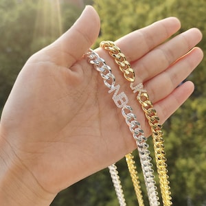 Könnte beinhalten: Silber- und goldfarbene Panzerketten-Halsketten mit Initialen-Anhängern. Die silberne Halskette hat die Buchstaben "NB" und die goldene Halskette die Buchstaben "JK". Die Ketten werden in einer Hand gehalten.