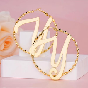 May include: A pair of gold-colored hoop earrings. Each earring features a large, cursive letter "Y" within a twisted rope-like frame. The earrings are displayed on a white surface against a soft pink background.