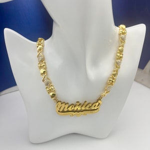 K&ouml;nnte beinhalten: Eine goldfarbene Halskette mit dem Namen "Monica" in kursiver Schrift. Die Halskette hat herzf&ouml;rmige und X-f&ouml;rmige Glieder mit kleinen, klaren Edelsteinen. Die Halskette wird auf einer wei&szlig;en Schaufensterpuppe pr&auml;sentiert.