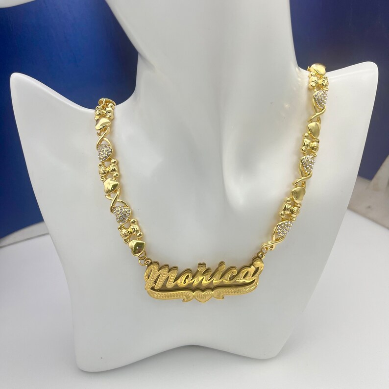K&ouml;nnte beinhalten: Eine goldfarbene Halskette mit dem Namen "Monica" in kursiver Schrift. Die Halskette hat herzf&ouml;rmige und X-f&ouml;rmige Glieder mit kleinen, klaren Edelsteinen. Die Halskette wird auf einer wei&szlig;en Schaufensterpuppe pr&auml;sentiert.