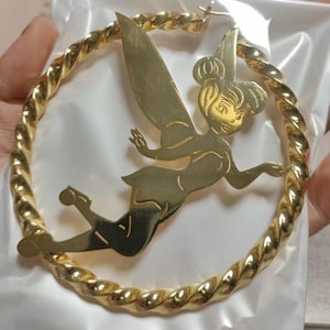May include: Gold-toned hoop earrings with a twisted rope design. The earrings feature a cutout of a fairy in flight. The fairy has large wings and is wearing a crown. The earrings are likely made of metal.
