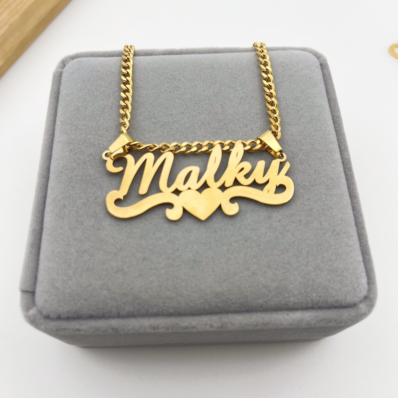 May include: Gold necklace with a cursive nameplate pendant that reads "Malky" with a small heart charm. The necklace is on a gray velvet box.