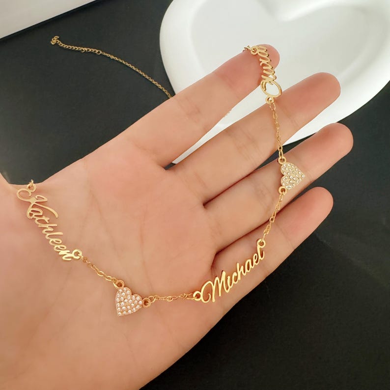 May include: Gold necklace with two cursive names, "Kathleen" and "Michael", separated by two heart-shaped charms with clear rhinestones.