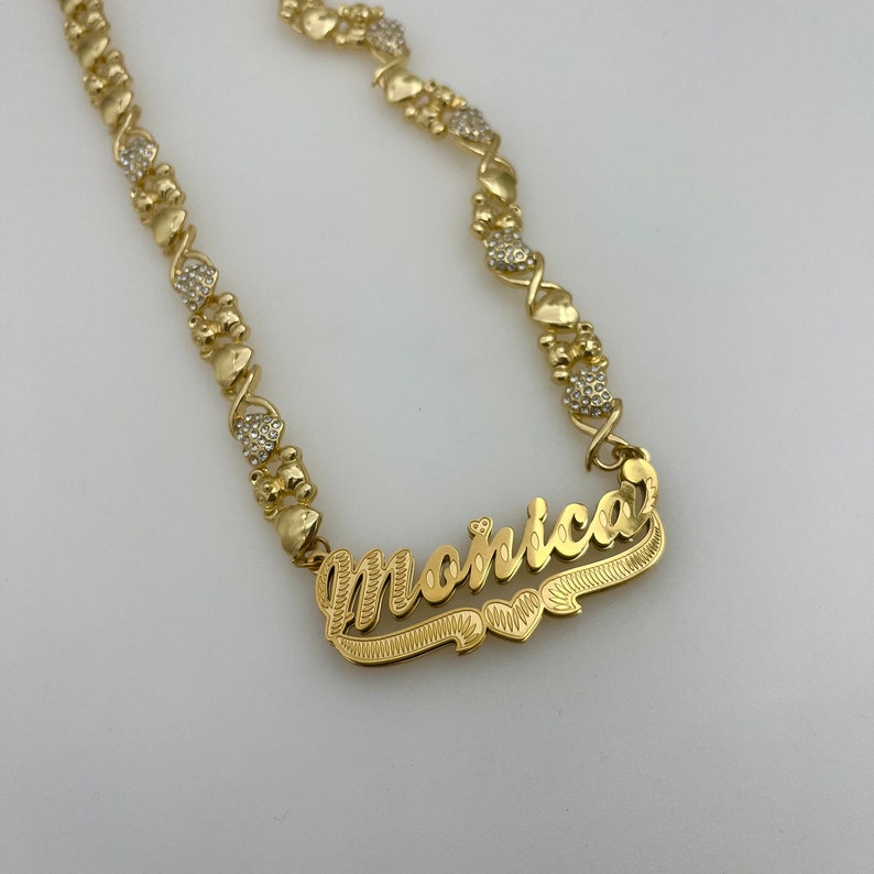 K&ouml;nnte beinhalten: Eine goldfarbene Halskette mit dem Namen "Monica" in Schreibschrift. Die Kette hat herzf&ouml;rmige und X-f&ouml;rmige Glieder, einige mit kleinen, klaren Edelsteinen. Das Namensschild hat einen dekorativen Rand.