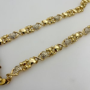 K&ouml;nnte beinhalten: Goldfarbene Halskette mit einem sich wiederholenden Muster aus Herzen, Teddyb&auml;ren und X's und O's. Die Halskette hat eine polierte Oberfl&auml;che, wobei einige Bereiche mit klaren Kristallen verziert sind. Das Design ist romantisch und verspielt.