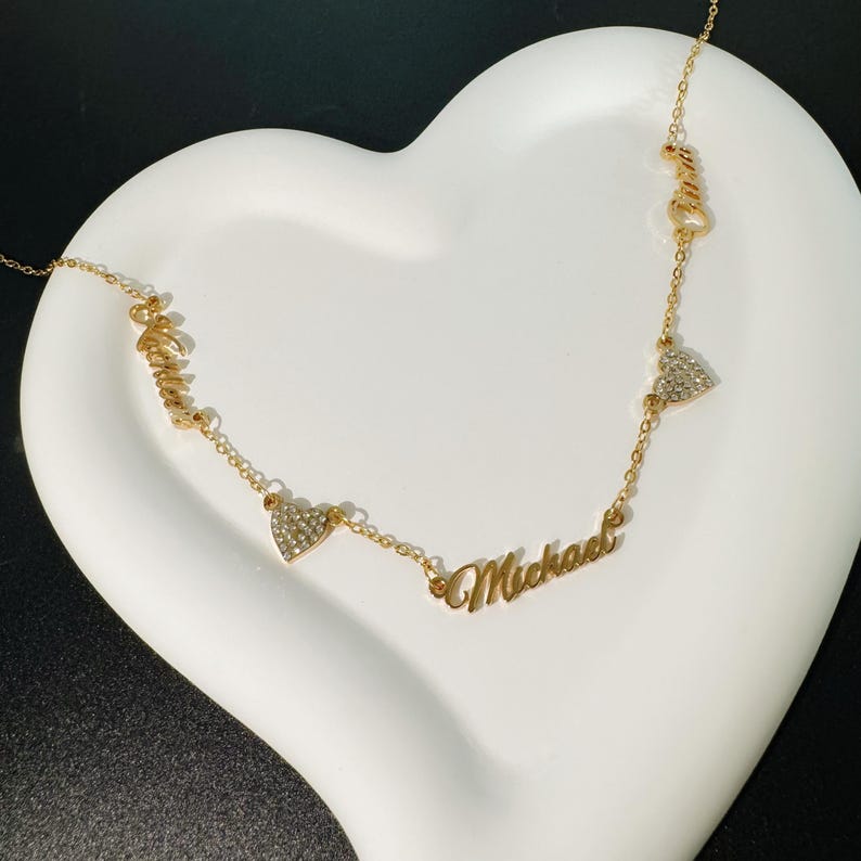 May include: Gold necklace with three heart charms and two names, "Amelia" and "Michael", spelled out in cursive.