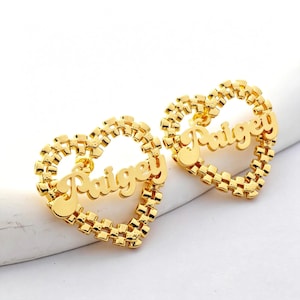 May include: A pair of gold-toned earrings featuring a heart-shaped frame with a textured, link-like border. Each earring has the name "Paigey" written in a cursive script font across the center of the open heart design, creating a personalized look.
