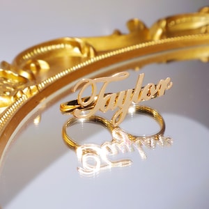 May include: Gold-colored hair clips with the name "Taylor" and "Love" in a cursive font. The clips are displayed on a reflective surface, with a gold-colored decorative frame in the background.