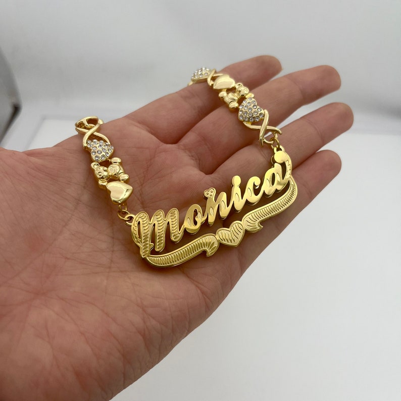 K&ouml;nnte beinhalten: Ein goldfarbenes Namensschildarmband mit dem Namen "Monica" in kursiver Schrift. Das Armband hat herzf&ouml;rmige und X-f&ouml;rmige Akzente mit eingebetteten, klaren Kristallen. Das Armband wird in einer Hand vor wei&szlig;em Hintergrund gehalten.