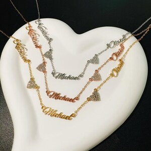 May include: Three gold, silver and rose gold necklaces with heart charms and personalised names, "Michael", "Olivia", and "Katherine", on a white heart-shaped surface.