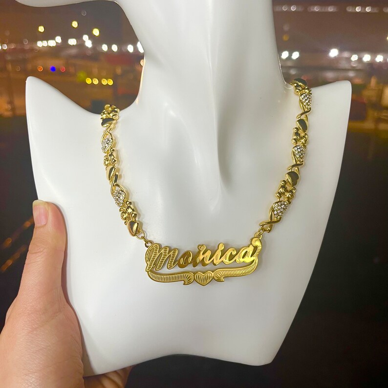 K&ouml;nnte beinhalten: Eine goldfarbene Halskette mit dem Namen "Monica" in Schreibschrift. Die Halskette hat eine Reihe von X- und O-Gliedern mit eingebetteten, klaren Kristallen. Das Namensschild ist zentriert und hat ein Herzdesign darunter.