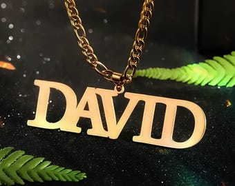 Puede incluir: Un collar dorado con el nombre "DAVID" en letras mayúsculas. El colgante cuelga de una cadena gruesa. El fondo es oscuro con efecto bokeh y acentos de helechos verdes.