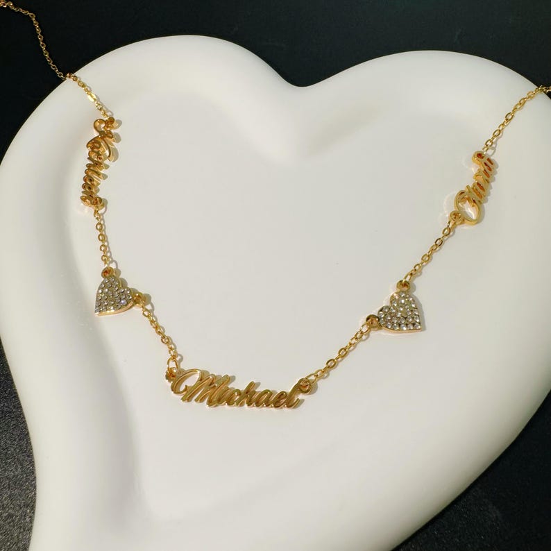 May include: Gold necklace featuring three names, "Olivia", "Michael", and "Olivia", written in a cursive script. Each name is separated by a small, clear heart-shaped charm with rhinestones.