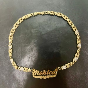 K&ouml;nnte beinhalten: Eine goldfarbene Halskette mit dem Namen "Monica" in kursiver Schrift. Die Halskette weist ein sich wiederholendes Muster aus Herzen und X's auf, wobei einige Abschnitte mit kleinen, klaren Kristallen akzentuiert sind. Die Halskette wird auf einer dunklen Oberfl&auml;che pr&auml;sentiert.