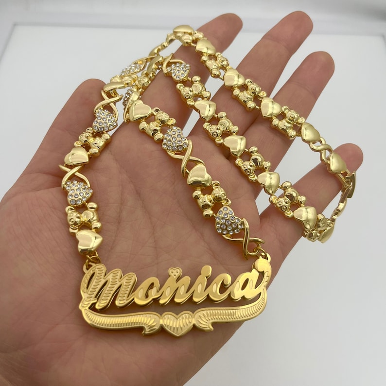 K&ouml;nnte beinhalten: Eine goldfarbene Halskette mit dem Namen "Monica" in Schreibschrift. Die Kette hat herzf&ouml;rmige und X-f&ouml;rmige Glieder, einige davon mit kleinen, klaren Kristallen. Die Halskette wird vor einem hellen Hintergrund pr&auml;sentiert.
