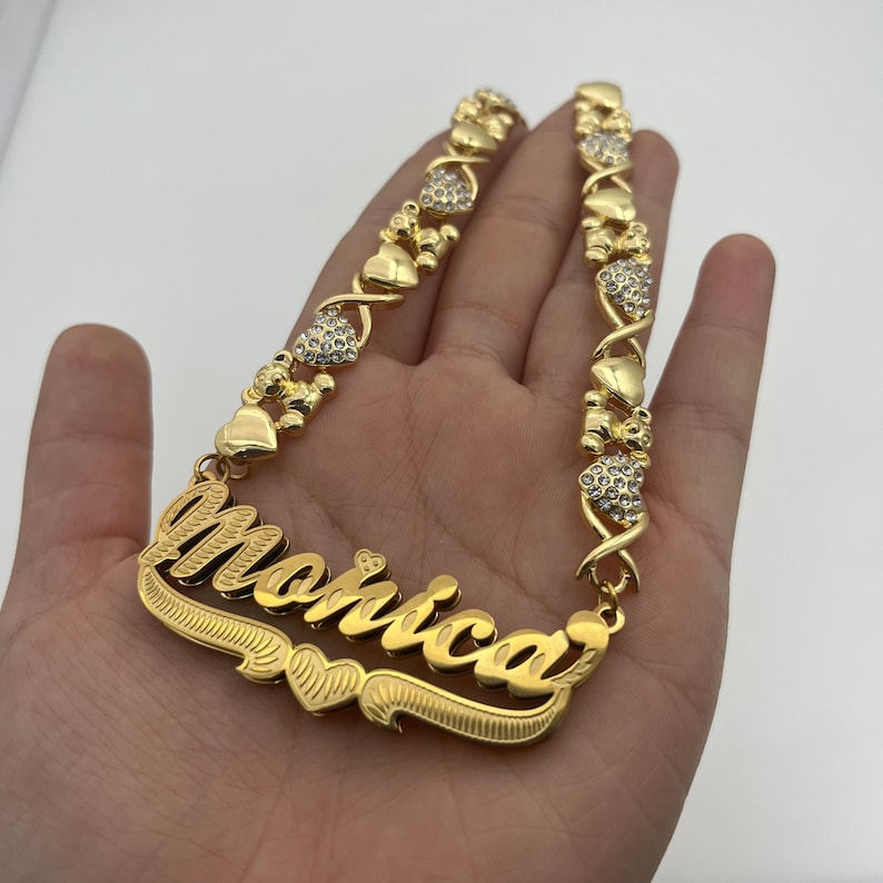 K&ouml;nnte beinhalten: Eine goldfarbene Halskette mit dem Namen "Monica" in Schreibschrift. Die Halskette hat herz- und b&auml;renf&ouml;rmige Akzente, einige davon mit kleinen, klaren Kristallen besetzt. Die Halskette wird in einer Hand vor wei&szlig;em Hintergrund gehalten.