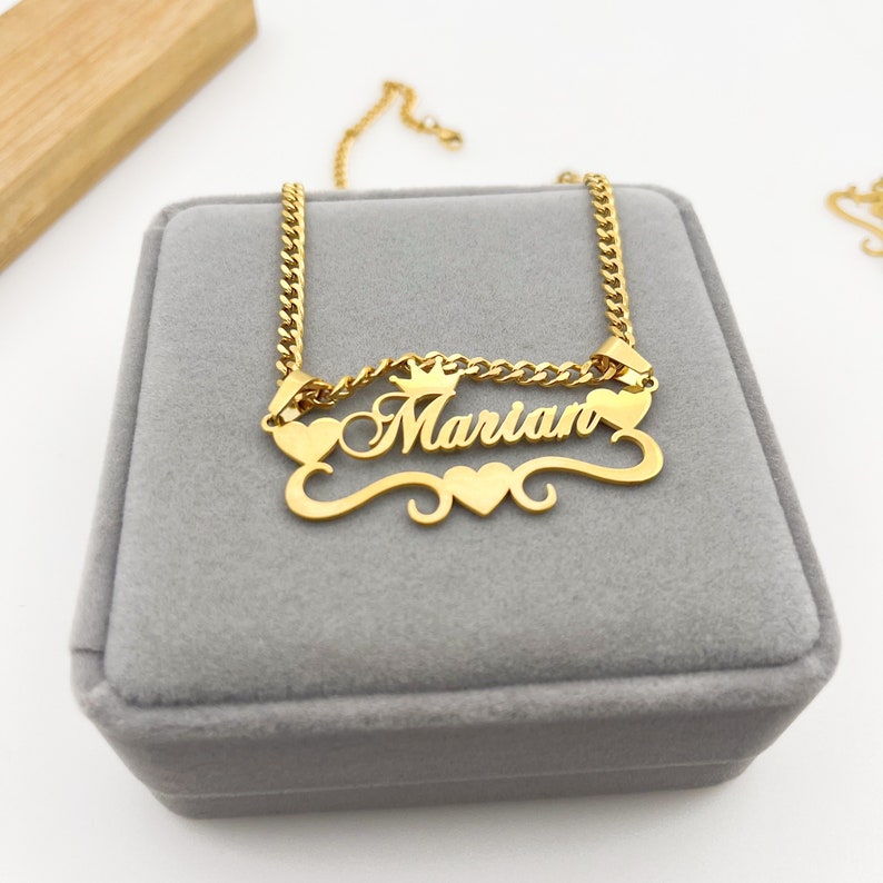 May include: Gold necklace with a cursive nameplate that reads "Marian" with a crown above the name and two hearts on either side. The necklace is in a gray velvet box.