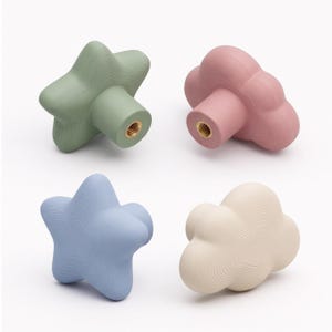 May include: A set of four cabinet knobs featuring two star-shaped and two cloud-shaped designs. The knobs are colored in sage green, dusty pink, light blue, and cream, each with a textured surface and a central brass-colored metal screw hole.