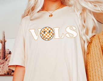 May include: A white t-shirt with the word "VOLS" in orange letters with a checkered smiley face in the "O". The model is wearing a brown cardigan.